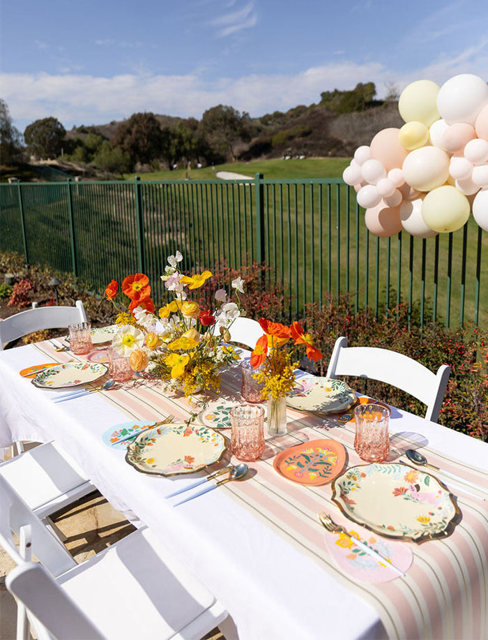 The Ultimate Easter Tablescape for 16: Folklore Easter by Sophistiplate LLC features designer paper plates, colorful glasses, wildflower centerpieces, white chairs, pastel balloons, and a scenic green fence leading to an open field.
