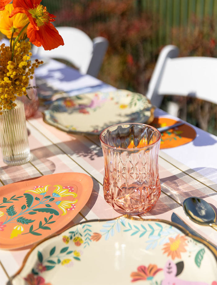 The Ultimate Easter Tablescape for 16: Folklore Easter by Sophistiplate LLC features designer plates, pink textured glassware, orange flowers in a vase, and white chairs on a pastel-patterned cloth—perfect for colorful outdoor celebrations.