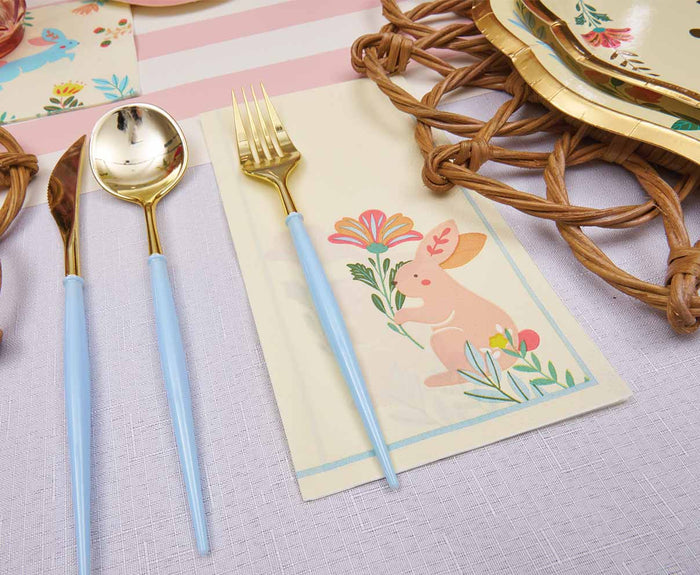 The Folklore Easter Table Setting by Sophistiplate features gold flatware with pastel blue handles on a bunny floral napkin, paired with designer disposable plates, a woven placemat, and a light pink-striped tablecloth for a charming look.