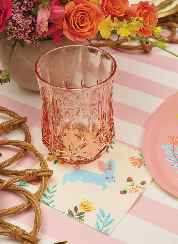 The Folklore Easter Table Setting by Sophistiplate features a pink glass on a floral napkin with a blue bunny, rattan coasters, designer plates, and a pink-and-white striped tablecloth—perfectly accented by a colorful flower arrangement.