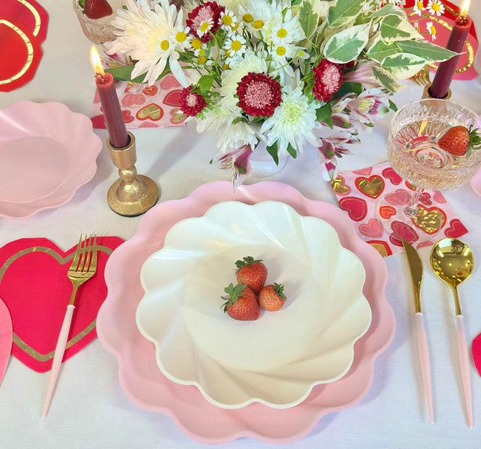 The Forever Blushing Table Setting by Sophistiplate includes pink and white designer plates with strawberries, gold and pink cutlery, heart placemats, red candles, a floral centerpiece, and a glass garnished with a strawberry.
