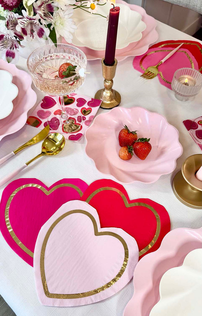 The Forever Blushing Table Setting by Sophistiplate includes heart-shaped disposable plates, gold utensils, pink napkins, a red candle, strawberries, and a glass with strawberry garnish on a floral-accented white tablecloth.