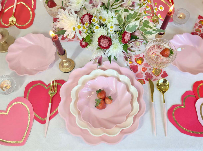 The Sophistiplate Forever Blushing Table Setting includes pink scalloped designer plates, gold cutlery, heart-shaped napkins and cards, red candles, and a white and pink floral bouquet for a festive tablescape-in-a-box.