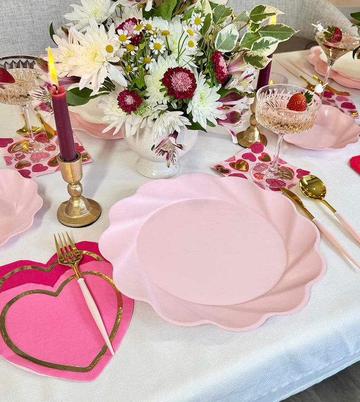 Sophistiplate's Forever Blushing Table Setting includes pink designer disposable plates, gold cutlery, crystal glasses with strawberries, heart-shaped napkins, and a floral centerpiece with white and pink blooms for a festive tablescape. Lit candles complete the look.