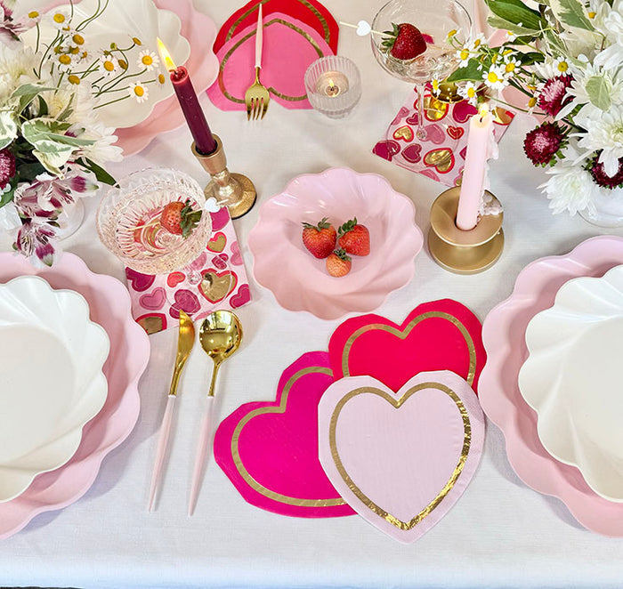 Create a romantic ambiance with the Forever Blushing Table Setting by Sophistiplate, featuring pink and white designer disposables, gold cutlery, heart-shaped placemats, fresh flowers, candles, and strawberries on a white tablecloth.
