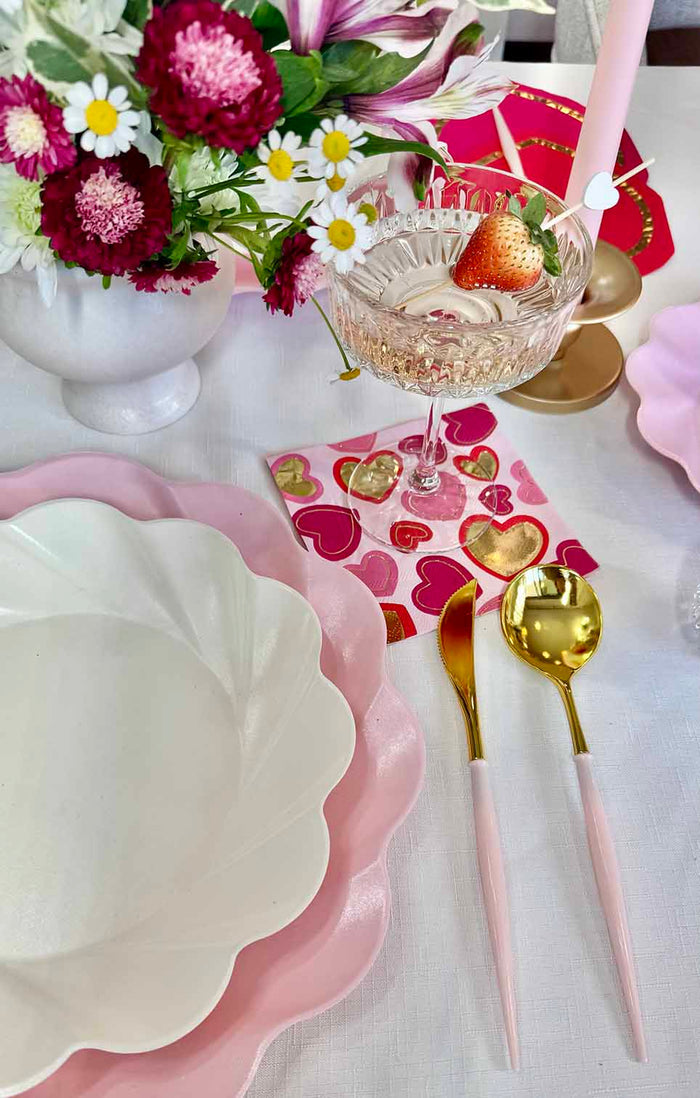 The Forever Blushing Table Setting by Sophistiplate includes pink scalloped plates, gold-pink cutlery, a strawberry-adorned pink cocktail glass, heart-print napkins, and vibrant flowers on a crisp white tablecloth.