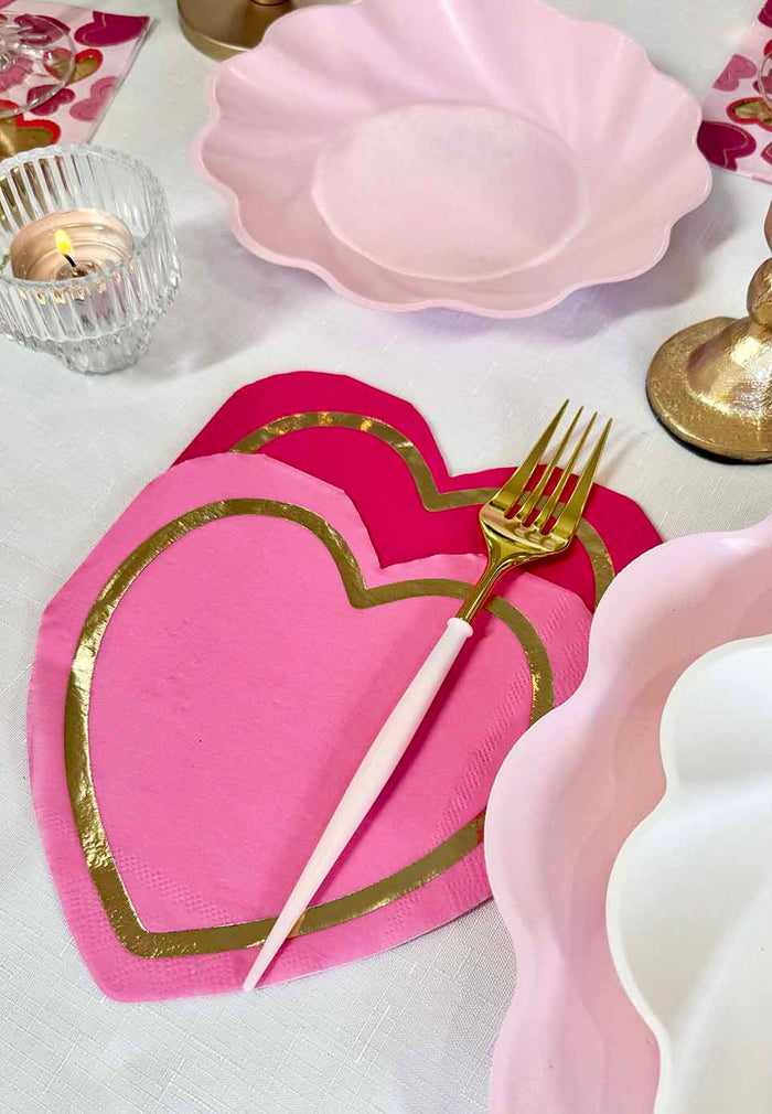 The Forever Blushing Table Setting by Sophistiplate features a gold and white fork on heart-shaped pink napkins with gold trim, designer disposable plates, a tealight candle, and gold candle holders atop a white tablecloth.