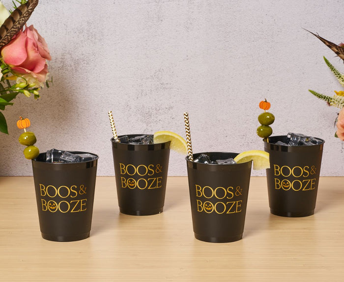 Four black Sophistiplate Frost Flex Cups "Boos & Booze" (8pk) are filled with ice and garnished with striped straws, lemon wedges, or skewered olives with pumpkin toppers; flowers appear in the background.