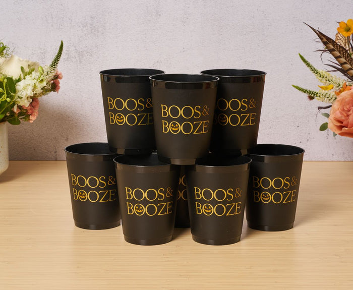 Eight black Frost Flex "Boos & Booze" cups by Sophistiplate are stacked on a wooden surface, surrounded by flowers. These reusable party cups add festive flair to your Halloween celebration.