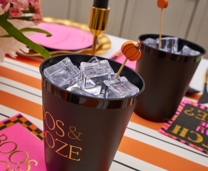 Close-up of Sophistiplate Frost Flex Cups "Boos & Booze"/8pk filled with ice and a stir stick on a colorful striped table, surrounded by pink napkins and festive tableware.