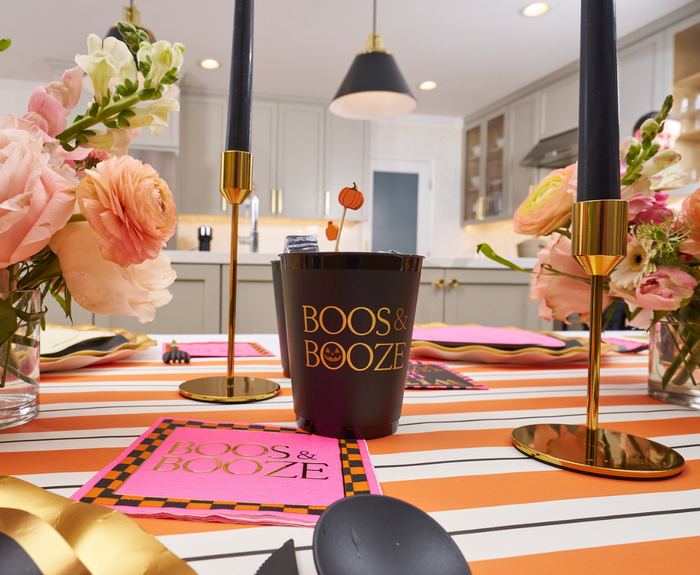 A festive table features black and gold candles, pink and orange flowers, a striped tablecloth, and a Sophistiplate Frost Flex Cup reading "Boos & Booze." These reusable Halloween cups (8pk) add flair to the bright, modern kitchen.