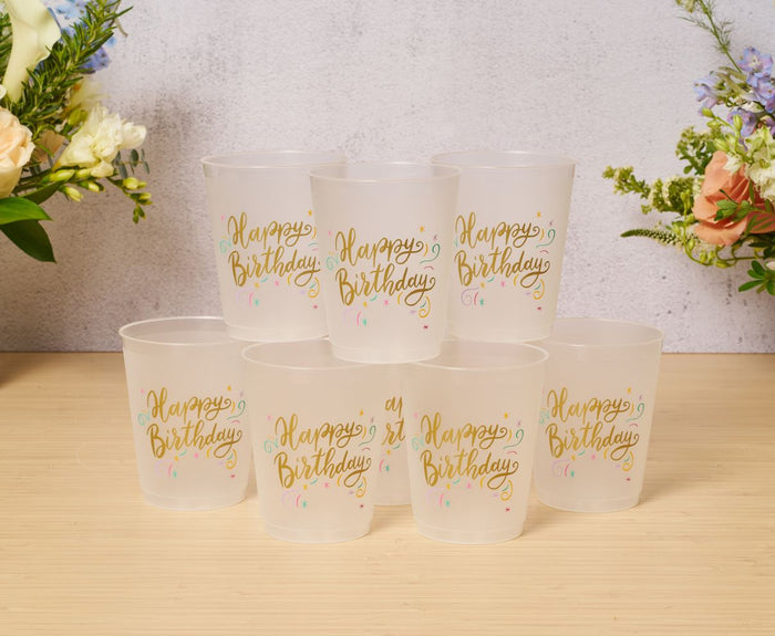 Eight reusable Frost Flex "Happy Birthday" cups by Sophistiplate, decorated with colorful confetti, are arranged in a pyramid on a wooden surface with floral arrangements visible in the background.