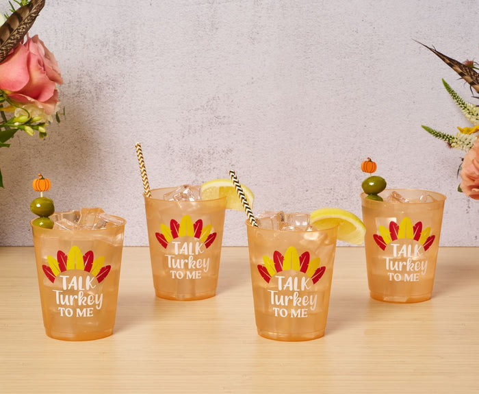 Four orange drinks in Sophistiplate Frost Flex Cups "Talk Turkey to Me" (8pk), topped with ice and fun garnishes—striped straws, olives, or a lemon wedge—sit on wood with bright flowers for a festive Thanksgiving vibe.