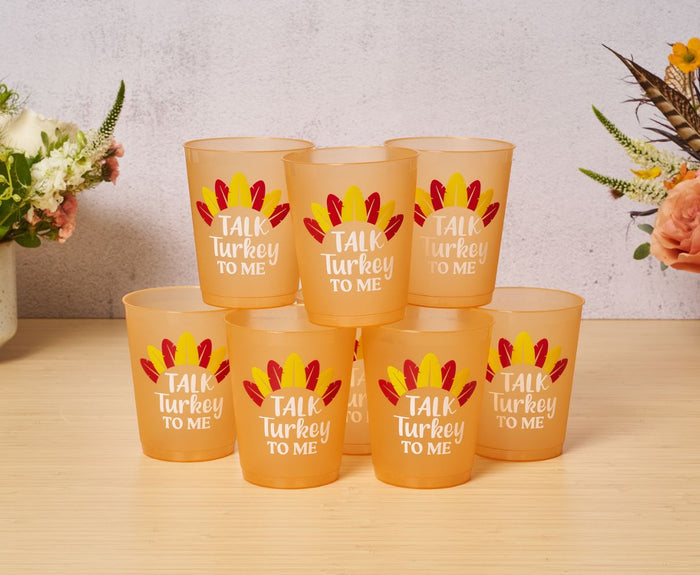 Eight orange Sophistiplate Frost Flex Cups, each with "Talk Turkey to Me" and feather designs, are stacked on a wooden table with flowers in the background. Perfect for Thanksgiving parties!.