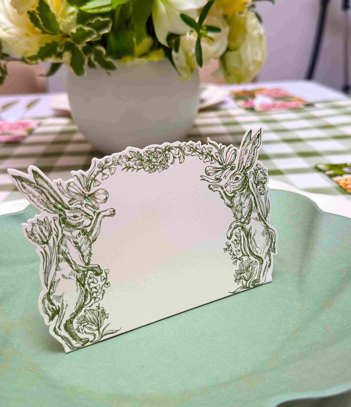 The Sophistiplate Garden Rose Table Setting features a decorative place card with rabbits and flowers on a pale green designer disposable plate, complemented by a vase of white blooms and a green-checkered tablecloth.