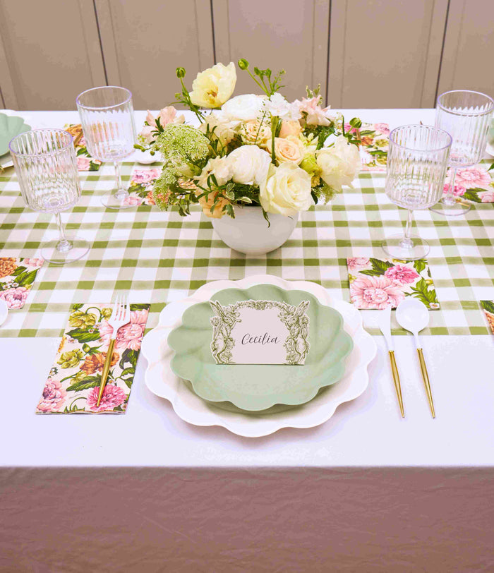 The Garden Rose Table Setting by Sophistiplate features green and white designer plates, gold cutlery, floral napkins, and a white and cream bouquet on a green checkered tablecloth. A card labeled Cecilia sits on one plate.
