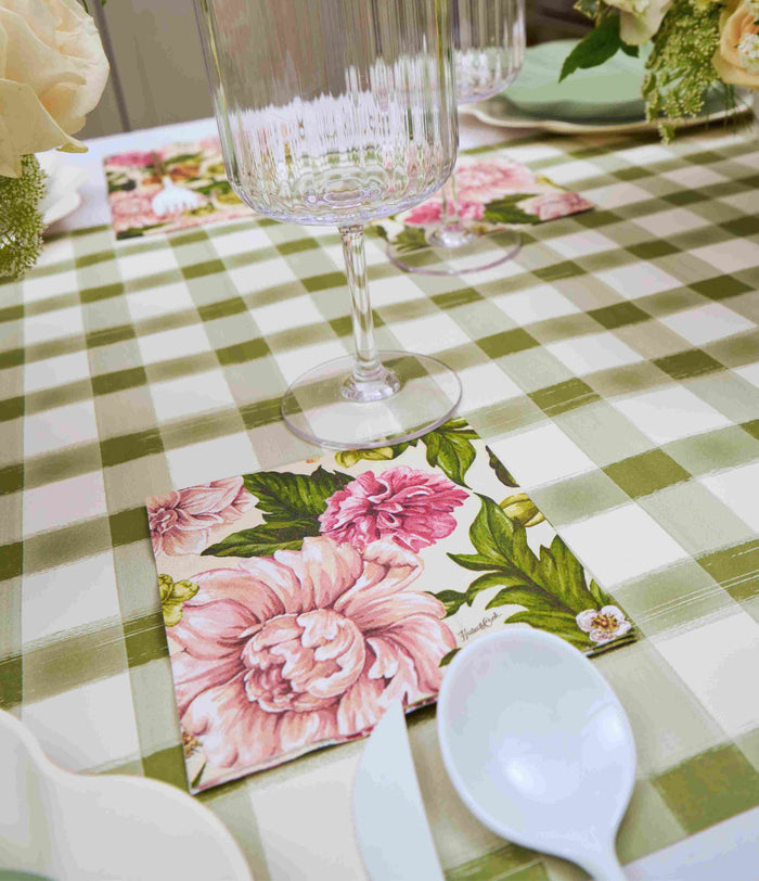 A close-up of the Sophistiplate Garden Rose Table Setting, featuring a green and white checkered tablecloth, floral napkin, clear glass, white spoon, and designer disposable plates adorned with white flowers.