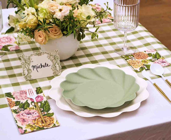 The Garden Rose Table Setting by Sophistiplate features designer floral disposable plates, gold cutlery, a wine glass, a floral arrangement, and a Michele name card on a green and white checked tablecloth.