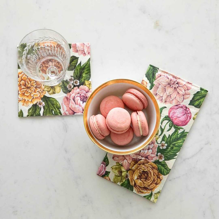 Pink macarons rest on a floral napkin beside a glass of water on Hester & Cook’s Garden Rose Cocktail Napkins (Pack of 20), all atop white marble—an elegant setup for spring gatherings.