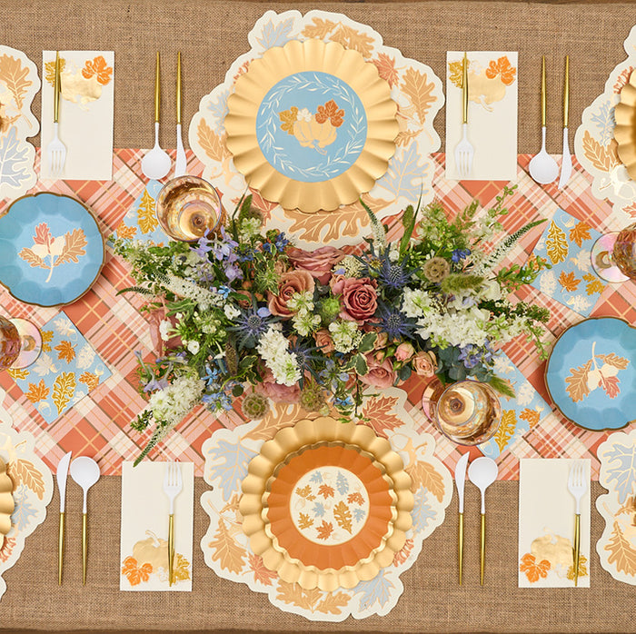 Create a stunning fall tablescape with Sophistiplate’s Gilded Harvest Table Setting—designer gold and blue plates with leaf motifs, plaid napkins, gold utensils, and floral centerpieces on a burlap tablecloth.