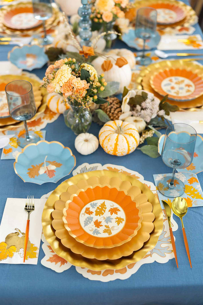 The Gilded Harvest Table Setting by Sophistiplate is a festive autumn party-in-a-box with gold, orange, and blue disposable plates, leaf napkins, gold flatware, blue glasses, mini pumpkins, fall flowers, and a blue tablecloth.