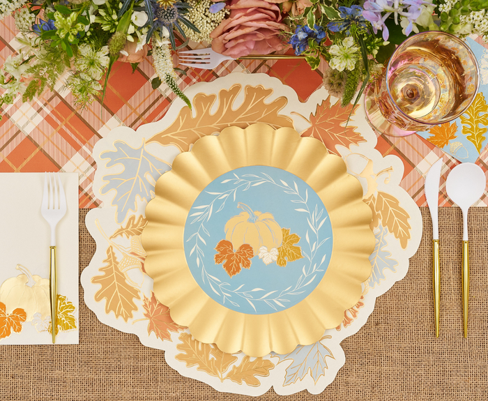 A festive autumn table features Sophistiplate's Gilded Harvest Dinner Plates (8pk) with pumpkin and leaves, set on a leaf placemat, surrounded by flowers, plaid napkins, gold utensils, and a glass of rosé.