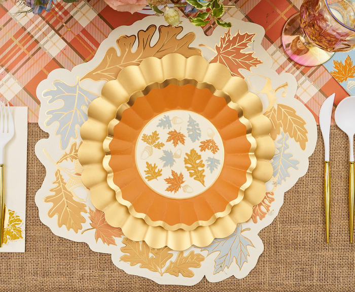 A festive autumn table features gold and orange scalloped leaf-print plates, gold utensils, a plaid runner, and Sophistiplate Gilded Harvest Die Cut Placemats (16ct).