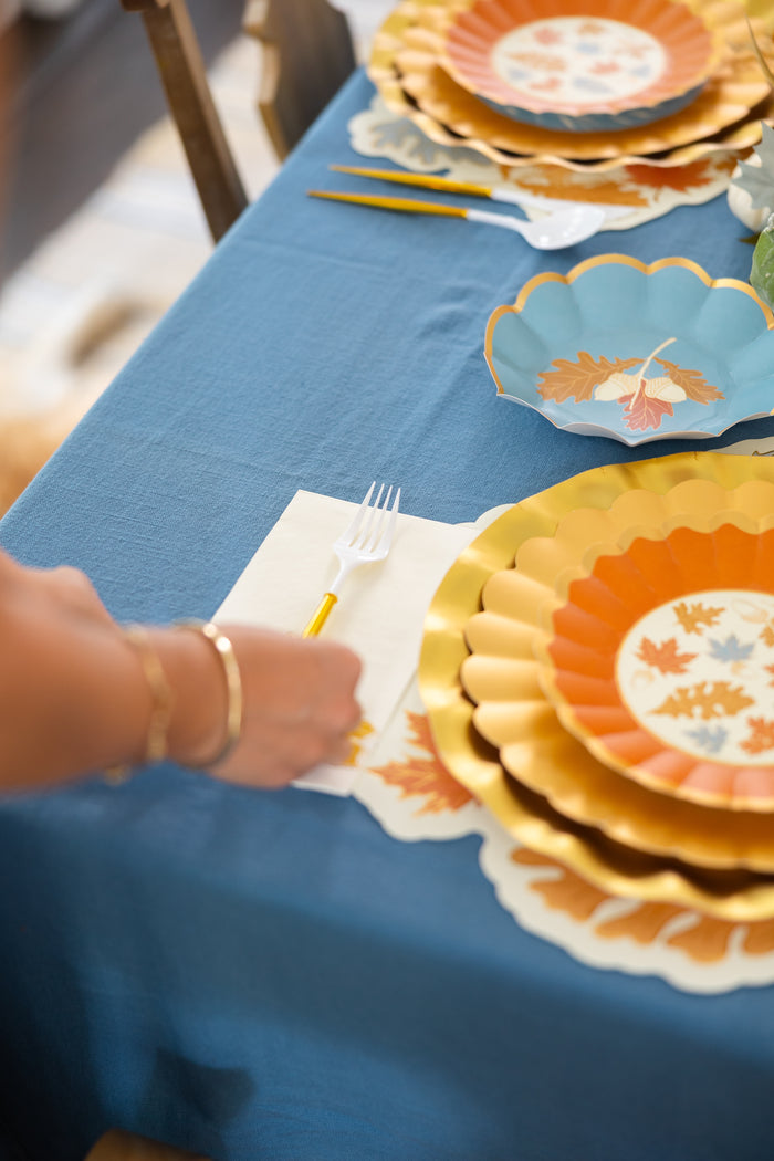 A close-up of a festive fall tablescape with Sophistiplate’s Gilded Harvest Table Setting—designer disposable plates featuring autumn leaf designs on a blue tablecloth—as a hand places a fork on a napkin beside the plates.