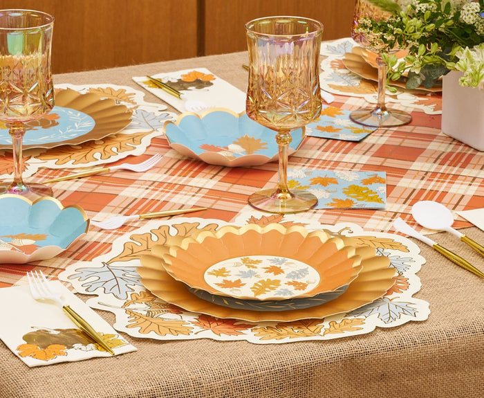 The Gilded Harvest Table Setting by Sophistiplate creates a chic autumn tablescape with designer disposable plates, gold cutlery, crystal glasses, napkins, a plaid tablecloth, and green plant centerpieces for an effortless party-in-a-box look.