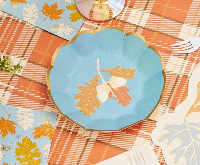 The Gilded Harvest Table Setting by Sophistiplate features a blue scalloped plate with gold trim and acorn motifs on an orange plaid tablecloth, bringing autumn charm alongside matching napkins and white plastic forks.