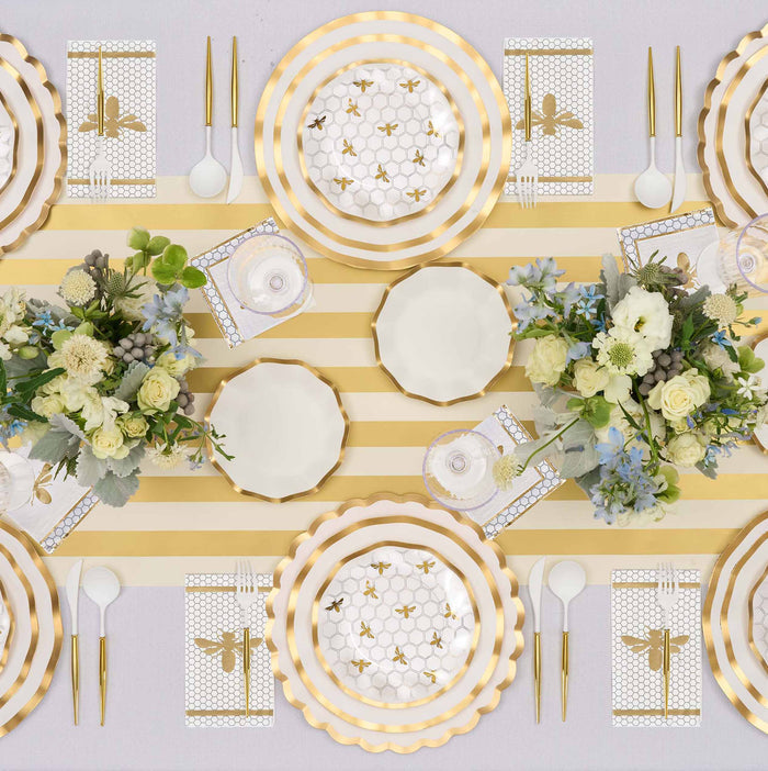 A tabletop set featuring Sophistiplate Honeybee Salad Plates (8pk), gold-striped paper plates with ruffled edges, bee-themed napkins, matching cups, and floral centerpieces, all elegantly displayed on a yellow and white striped table runner.