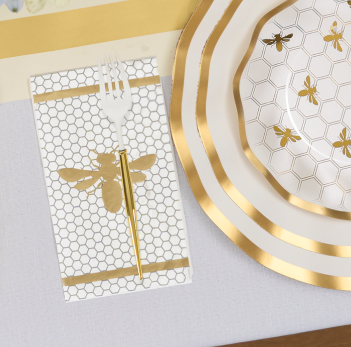 The Gilded Honey Table Setting by Sophistiplate LLC features designer disposable hexagon bee-patterned plates, a matching bee napkin, a white fork, and a gold-handled knife for an elegant white and gold table décor.