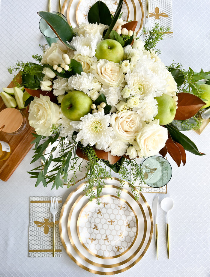 The Gilded Honey Table Setting by Sophistiplate LLC features gold and white designer plates, gold cutlery, bee-themed napkins, and a centerpiece of white flowers, greenery, and green apples for an elegant, fresh look.