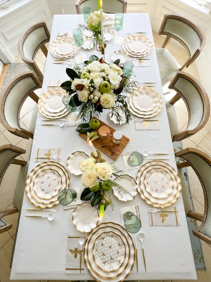 The Gilded Honey Table Setting by Sophistiplate LLC features a designer six-person ensemble with gold and white disposable plates, white napkins, green glasses, floral centerpieces, candles, and gold bee placemats for a sophisticated celebration.