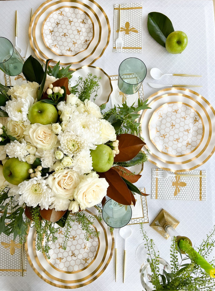 The Gilded Honey Table Setting by Sophistiplate LLC features gold and white designer plates, gold cutlery, green apples, tinted glasses, a lush centerpiece, and bee-themed napkins—perfect for stylish gatherings.