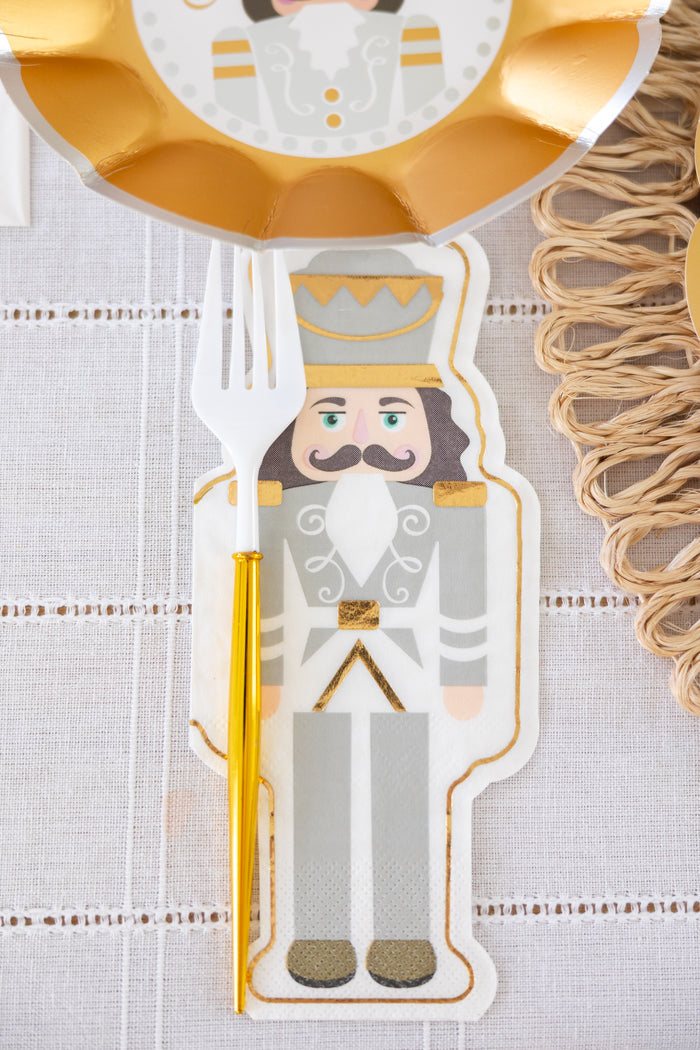 A Sophistiplate Gilded Nutcracker Guest Towel (16/pkg) with a gray, gold, and white nutcracker soldier design is set on the table. A white plastic fork with a gold handle rests atop, beside a gold plate—perfect for holiday gatherings.