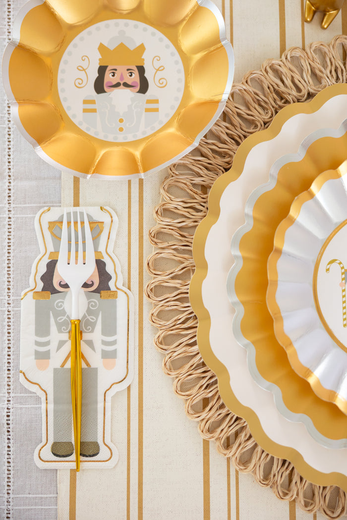 A gold and white party table is set with scalloped plates, a gold-rimmed nutcracker design plate, and a fork atop Sophistiplate's Gilded Nutcracker Guest Towel napkins (16/pkg), all on a striped tablecloth with woven placemats.