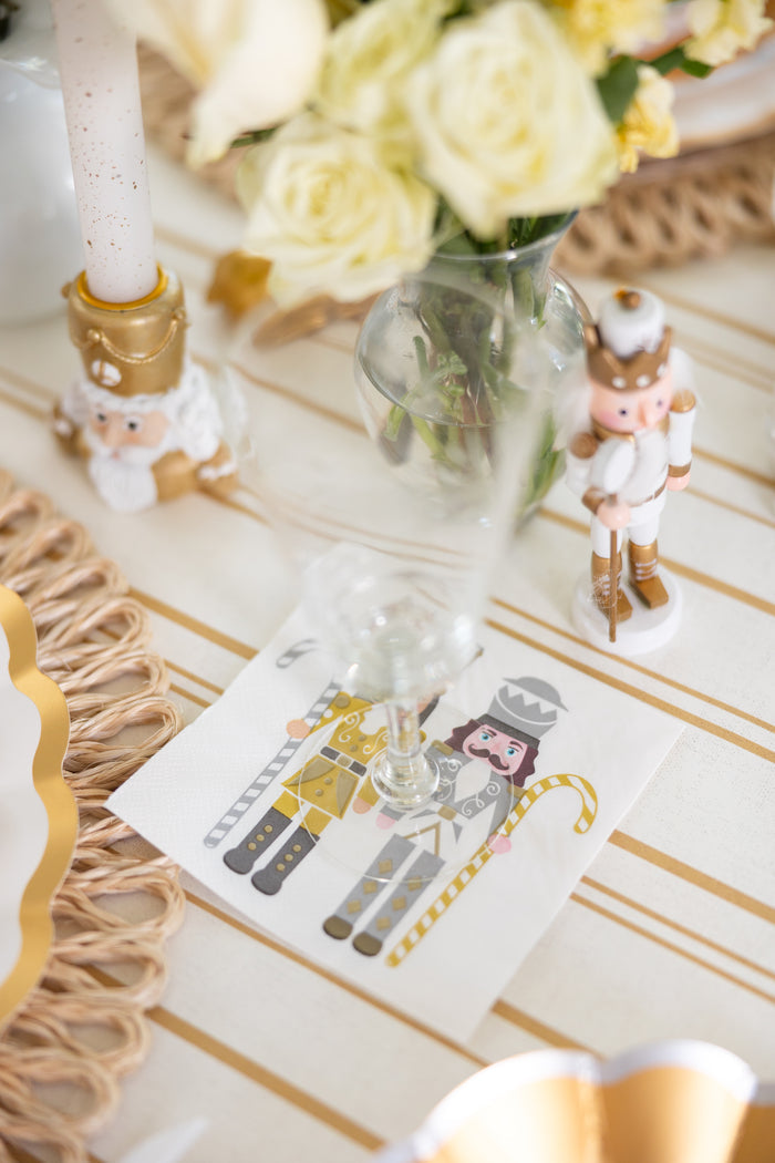 A chic table setup includes Sophistiplate’s Gilded Nutcracker Paper Cocktail Napkins beneath a wine glass, accented by nutcracker figurines, white roses, and gold holiday decor on a white and gold striped tablecloth.