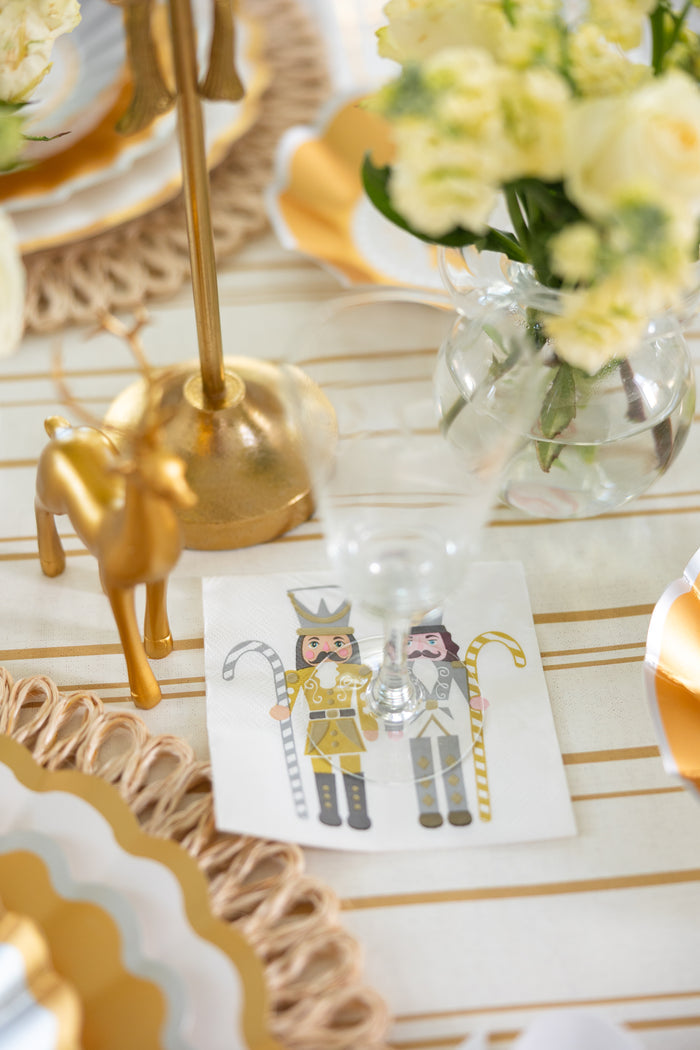 A festive table features a gold reindeer figurine, a clear glass, white and yellow flowers in a vase, and Sophistiplate's Gilded Nutcracker Paper Cocktail Napkins (20pk) with two nutcrackers—perfect for your holiday decor.