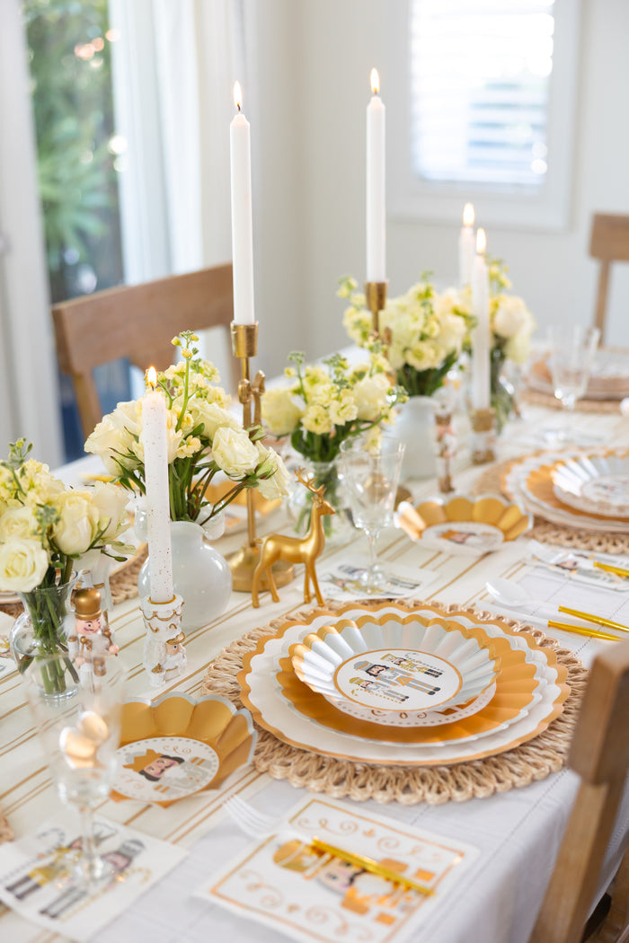A bright, elegant dining table is set with gold-rimmed plates, white and yellow flowers, tall candles, decorative menus, and Sophistiplate’s Gilded Nutcracker Paper Lunch Napkins for a fresh and inviting holiday look.