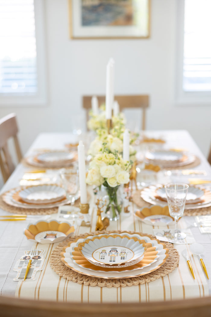 A stylish dining table set featuring Sophistiplate’s Gilded Nutcracker Dinner Plates (8-pack), gold cutlery, white napkins, tall white candles, and a centerpiece of white and yellow flowers in a bright, sunlit room.