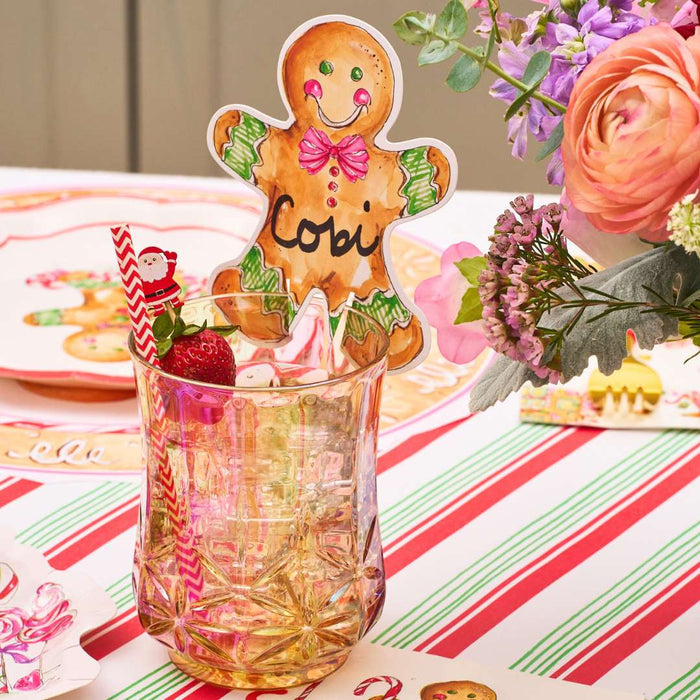 A festive table setting features a Rosanne Beck Gingerbread Cookie Cup Place Card (10pk) as a charming Christmas name card, paired with a drink, Santa straw decoration, strawberries, colorful flowers, and striped placemats.