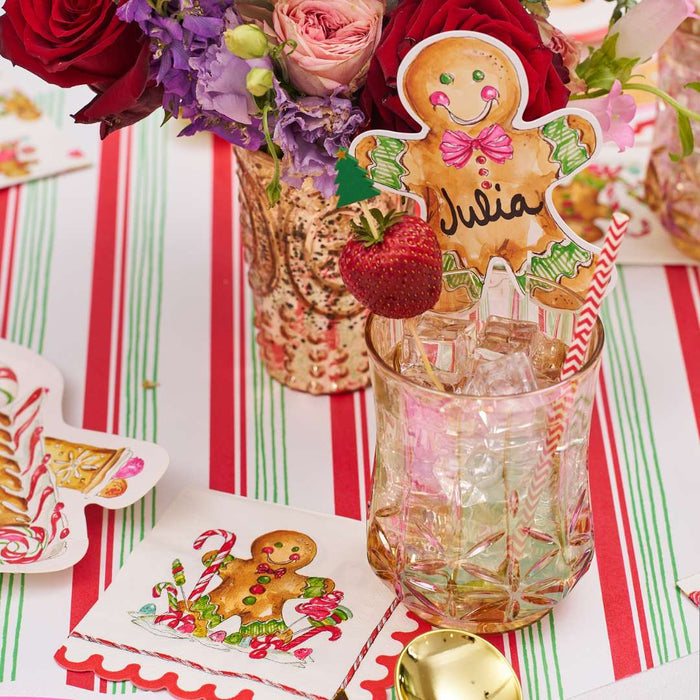 A festive table features Rosanne Beck's Gingerbread Cookie Cup Place Card/10pk, a striped red, green, and white tablecloth, holiday name cards, a floral centerpiece, and napkins with gingerbread and candy cane designs.