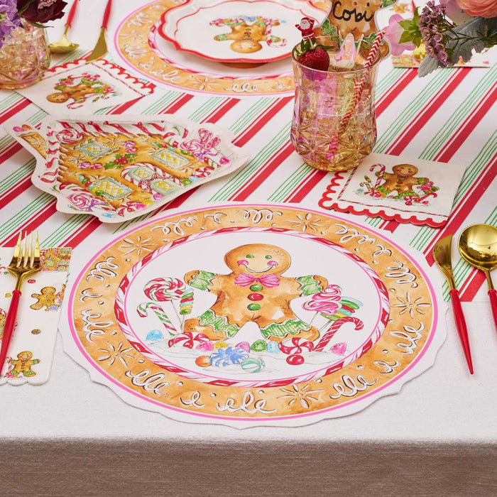 A festive Christmas table is set with Rosanne Beck Gingerbread Cookie Round Placemats (10pk), gingerbread and candy cane plates, gold flatware, a striped holiday tablecloth, and floral decor for a cheerful holiday setting.