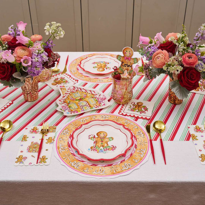 A festive table features gingerbread-themed plates, pink and red floral centerpieces, gold cutlery, gingerbread house decor, and the Rosanne Beck Red & Green Striped Table Runner (20x12") for a charming Christmas look.