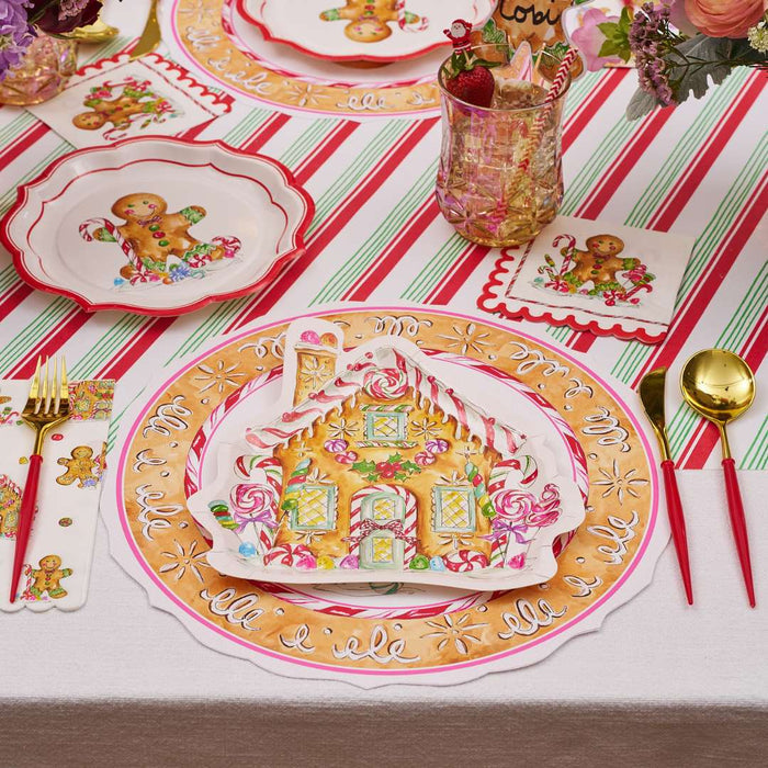 A festive table setting with gingerbread house-themed plates, gold cutlery, pink glasses, floral napkins, and the Rosanne Beck Red & Green Striped Table Runner (20x12") brings holiday cheer to your celebration.