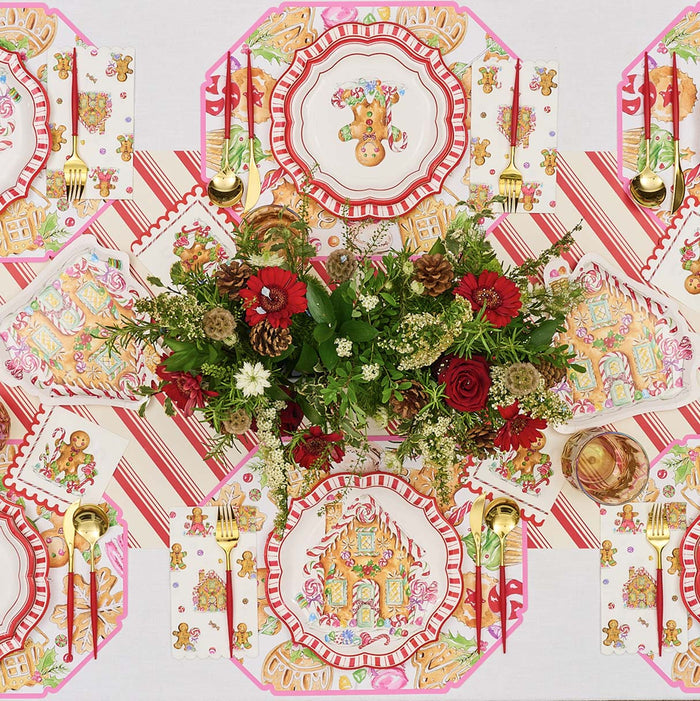 Create a festive table with the Gingerbread: Christmas Table for 8 set, featuring gingerbread-themed disposable plates, striped napkins, gold cutlery, and a red-and-white floral centerpiece on a holiday-patterned white cloth.