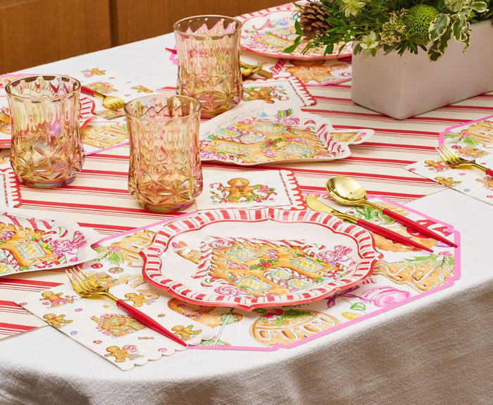 Create a charming Christmas table with the Gingerbread Table Setting by Sophistiplate, featuring designer paper plates, gold utensils, pink and gold glasses, and a festive tablecloth accented with a white vase of greenery.