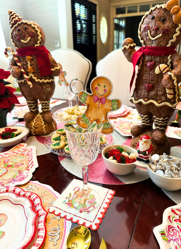 Create a festive look with the Sophistiplate Gingerbread Table Setting, featuring two large gingerbread figures, a gingerbread cookie in a glass, designer plates, candies, and holiday napkins on a wooden table.