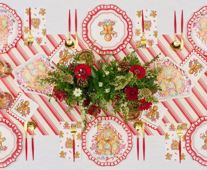 Create a festive tablescape with the Sophistiplate Gingerbread Table Setting, featuring red and white striped placemats, gingerbread-themed disposable plates, gold cutlery, and a red-and-white floral centerpiece with greenery.